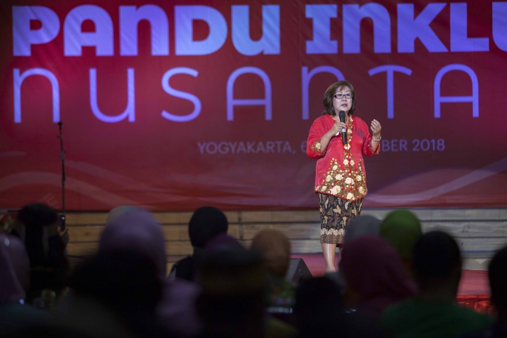 Perempuan dari komunitas Cina Benteng menggunakan kebaya merah dan kain batik, berkacamata sedang di atas panggung menggegam mikrofon disaksikan oleh banyak orang sebagai penonton. Sebagai latar belakang, terdapat tulisan Pandu Inklusi Nusantara